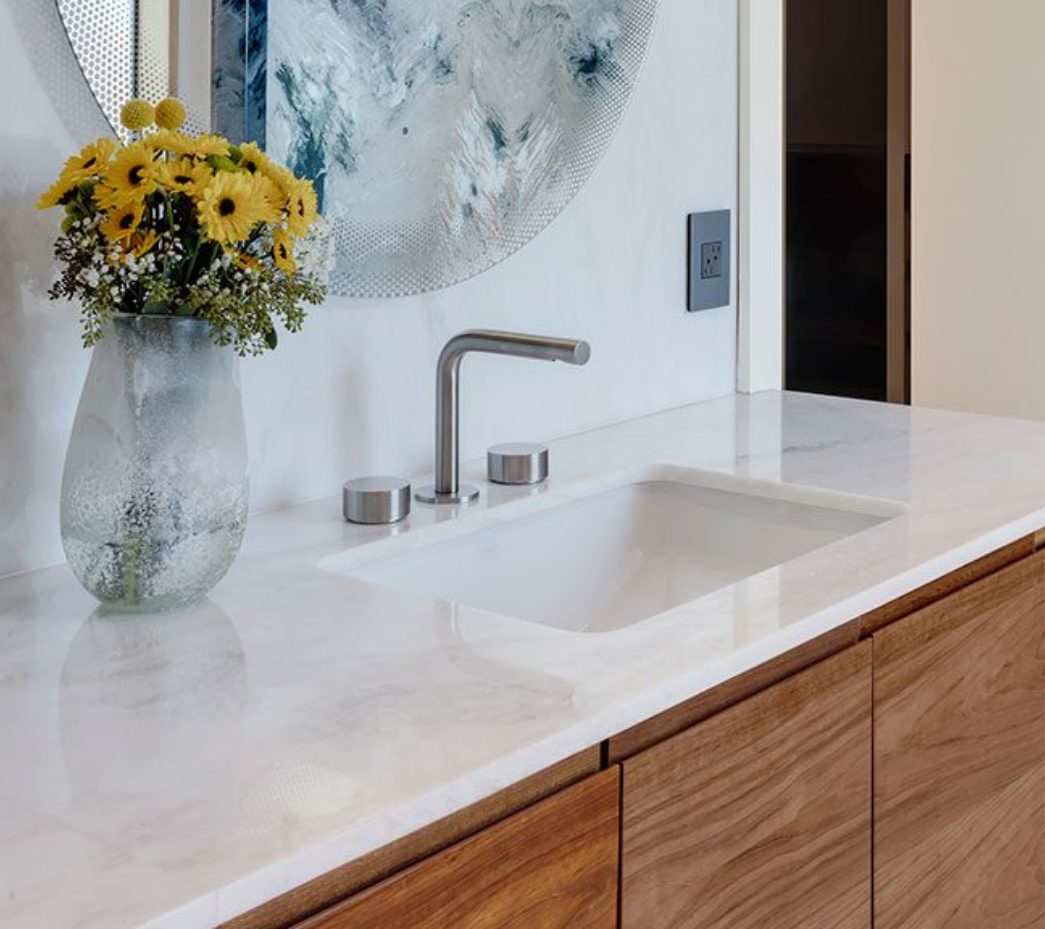 about-img-1 Modern bathroom sink with a chrome faucet on a marble countertop, wooden cabinets below, and a vase of yellow and white flowers next to a round, textured mirror.