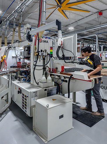 Edgebender A person operating a large industrial machine in a factory setting, surrounded by panels and machinery, with overhead pipes and lighting visible.