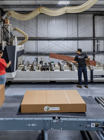 CNC2(3) Two workers operate a large industrial machine in a factory, with cardboard boxes on a conveyor belt in the foreground.