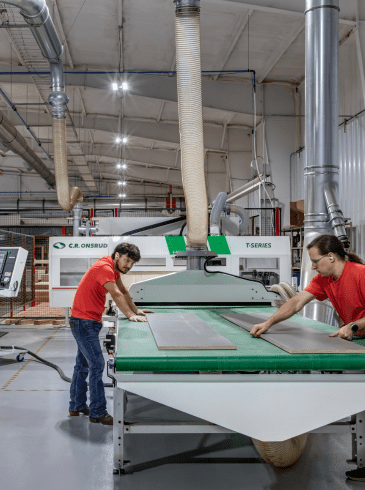 fix-image2(3) Three people work in a factory, operating machinery and handling large panels on an assembly line in a brightly lit industrial setting.