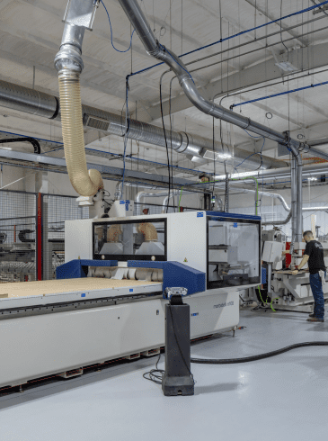 p2 Two people operate large woodworking machinery in a clean, industrial facility with metal ducts and safety barriers.