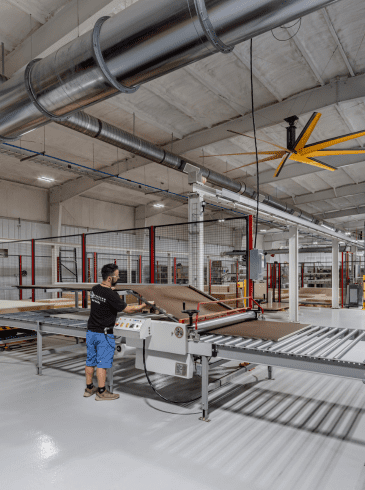 p6 A worker operates a machine to process large sheets of material in a spacious, modern factory with exposed ductwork and a yellow ceiling fan.