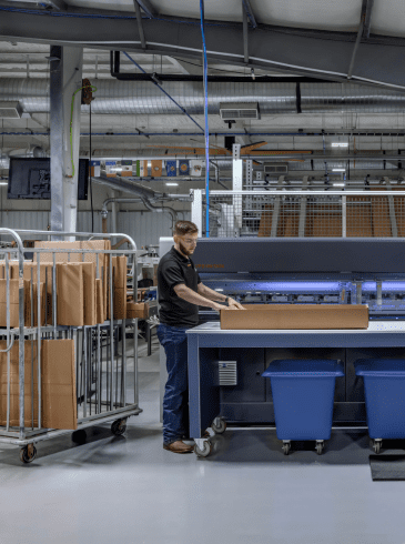 packaging(2) Two people operate a large packaging machine in a factory, assembling cardboard boxes with carts, bins, and computer monitors nearby.